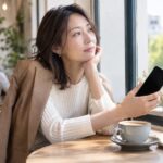 窓辺でスマートフォンを手に考える女性